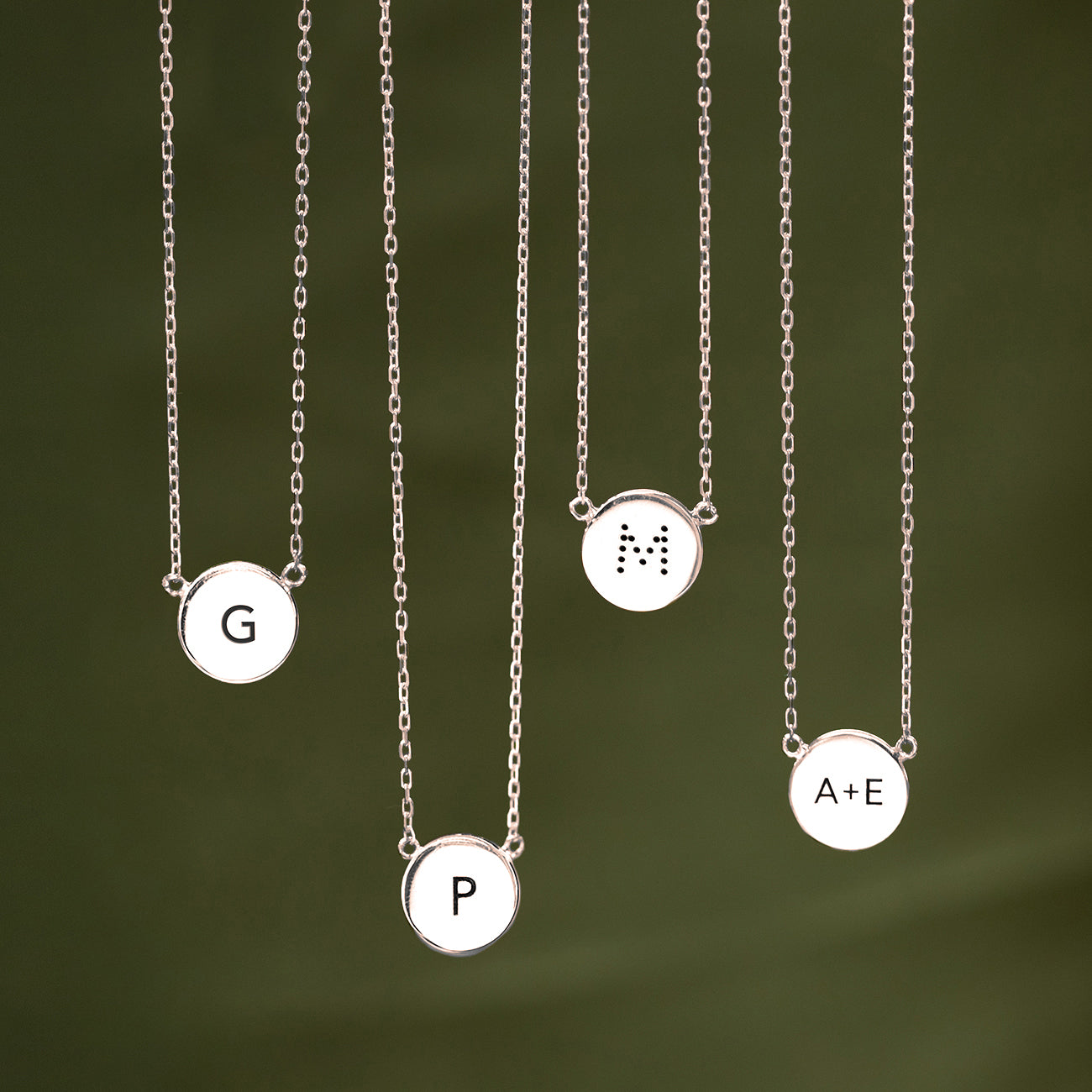 Four sterling silver engravable disc necklaces engraved with initials, on a green background