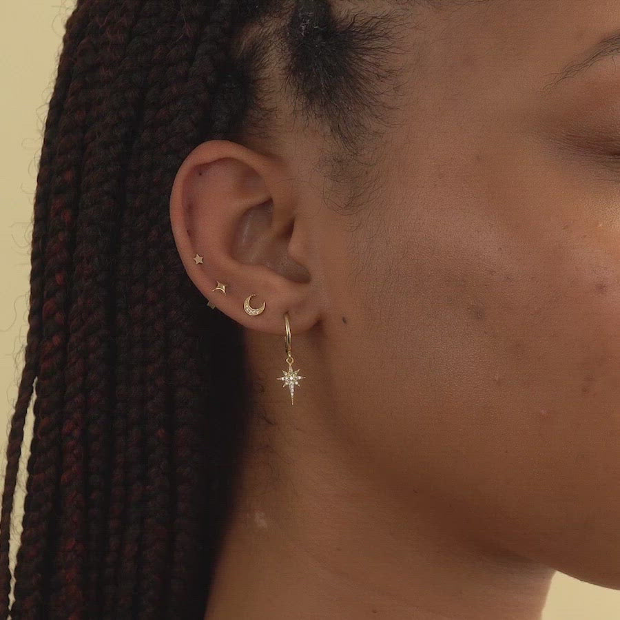 Woman wearing gold starburst huggie earring, gold crescent moon pave stud, gold flare star single stud, and gold tiny star single stud 