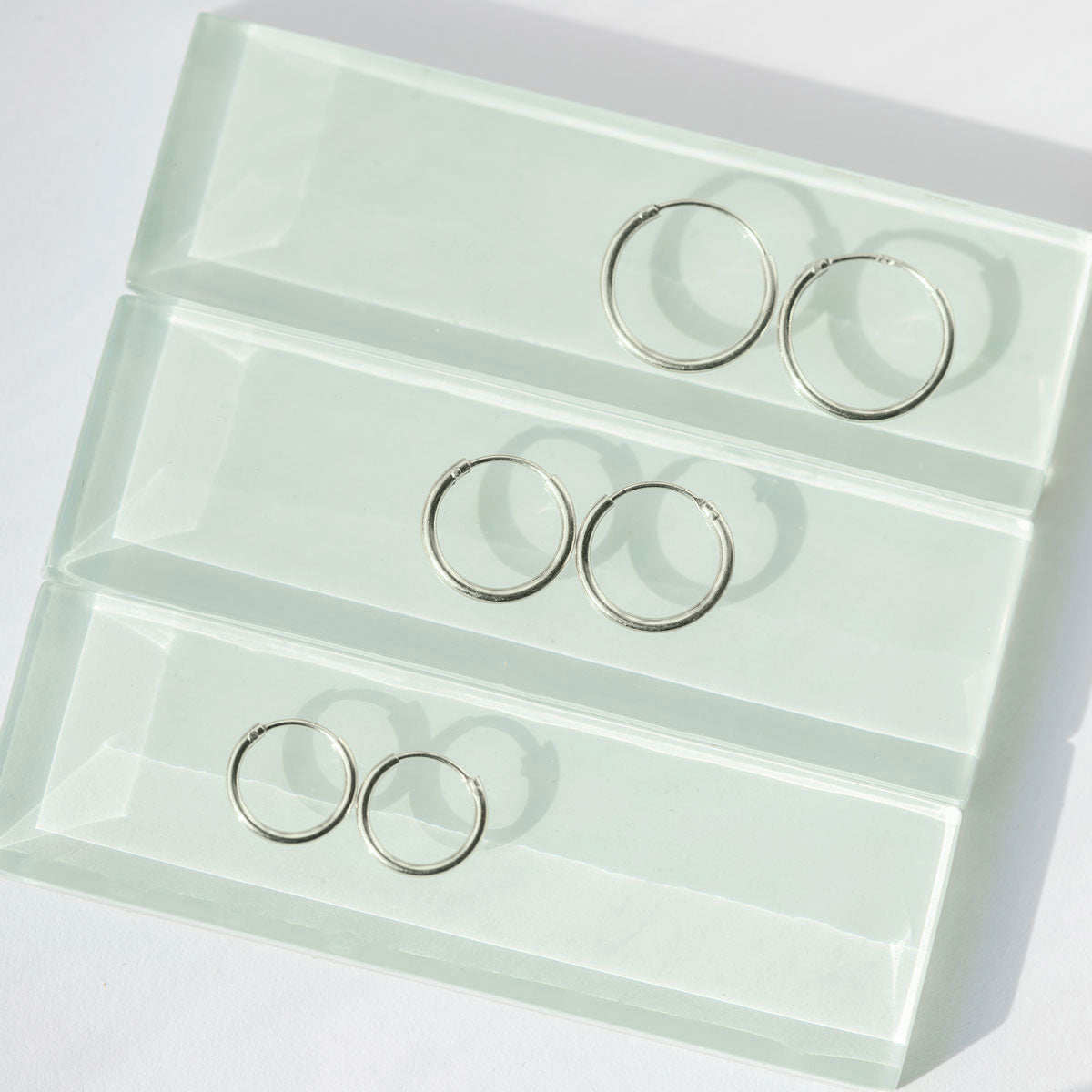 Sterling silver thin huggie hoops in three sizes laid on pale green glass tiles