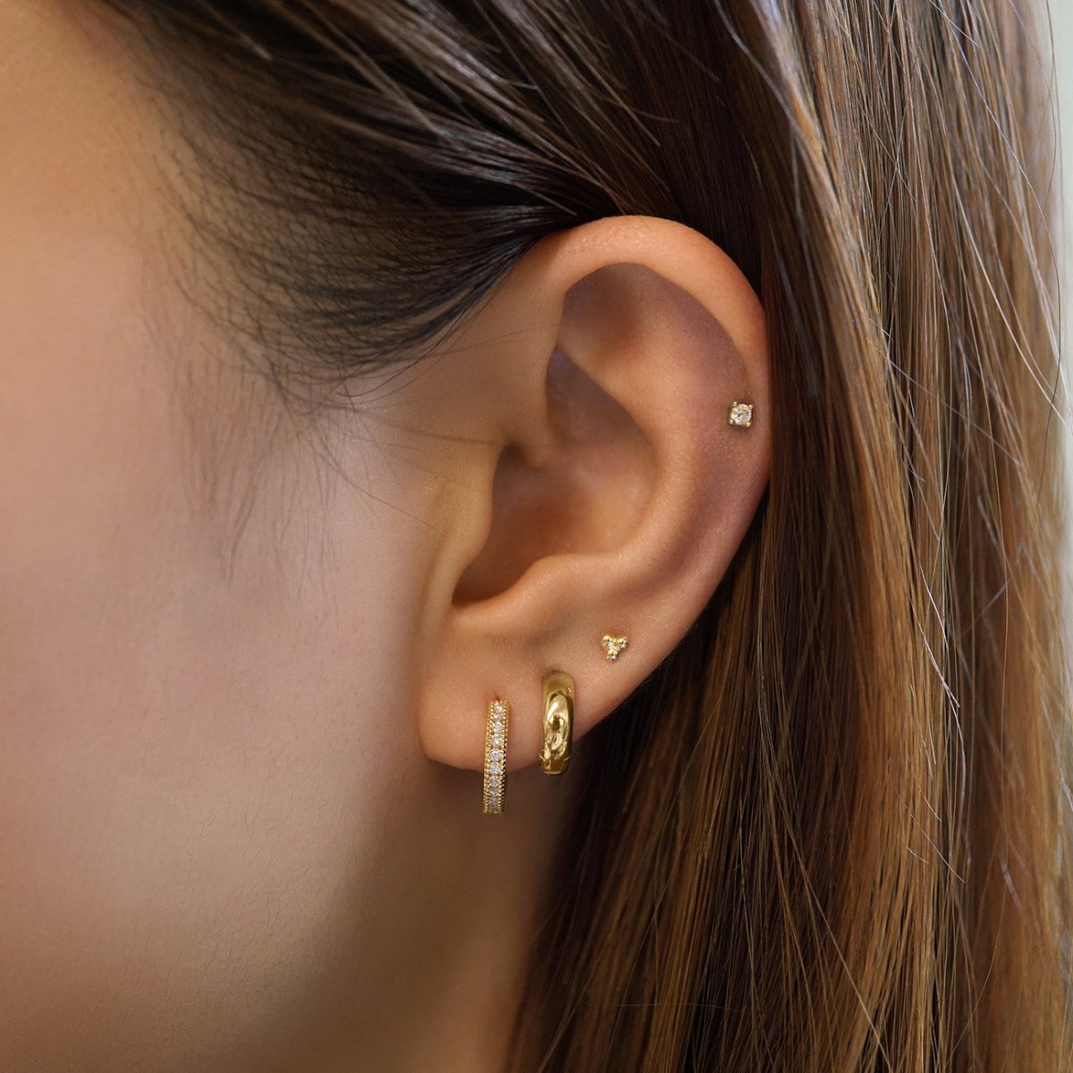 14K gold milgrain diamond huggie shown on a model's lobe with a trio stud and helix stud