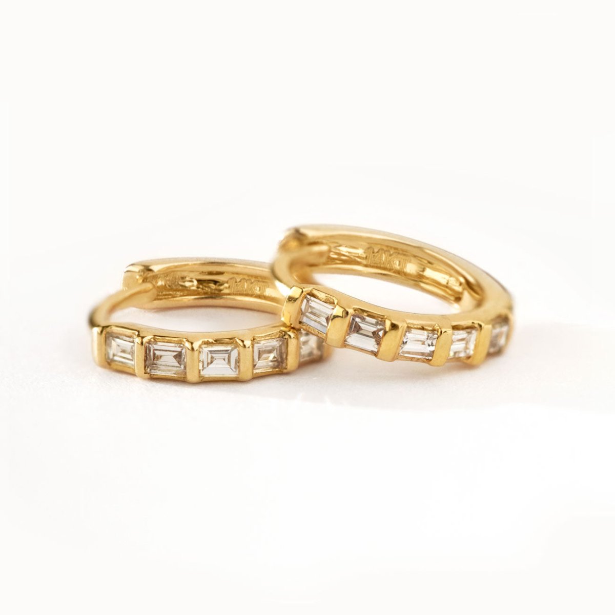 Pair of 14K gold baguette diamond huggie hoops shown resting on a white surface