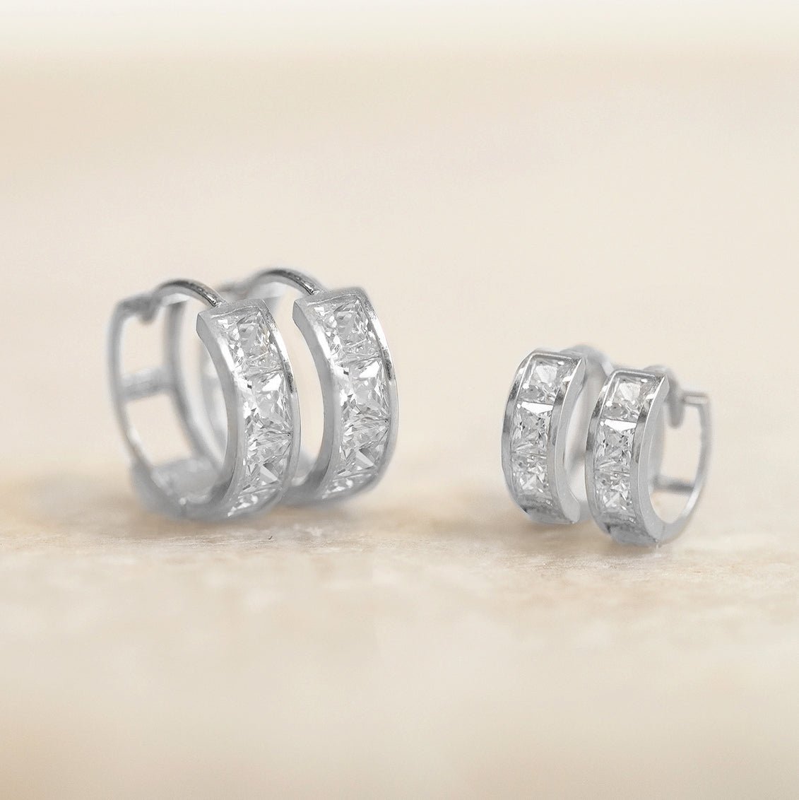 Two sizes of sterling silver white zirconia huggie hoops shown side by side on a cream background