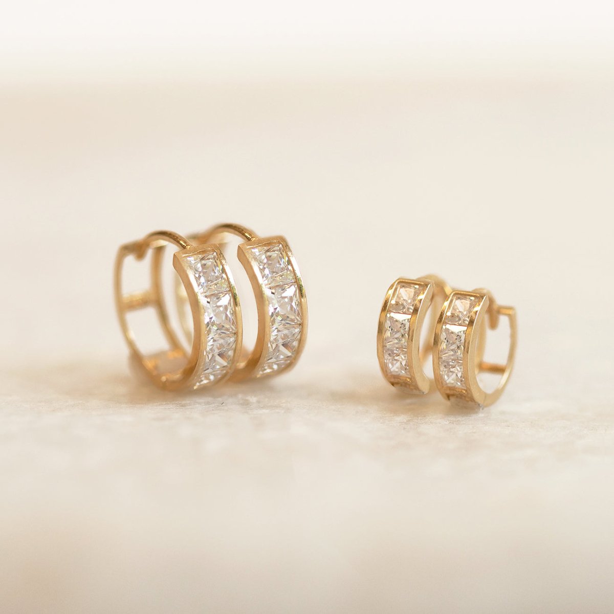 Two sizes of gold vermeil princess-cut white zirconia huggie hoops shown on a cream background